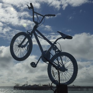 A blue painted BMX leaps from the top of a disused telegraph pole, against a blue sky with puffy white clouds, and with Brighton Pier in the background 