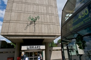Two green bikes leap out of the facade of a block of flats, above the entrance to Roehampton Library.  In the foreground stands a bus signposted 'Roehampton'