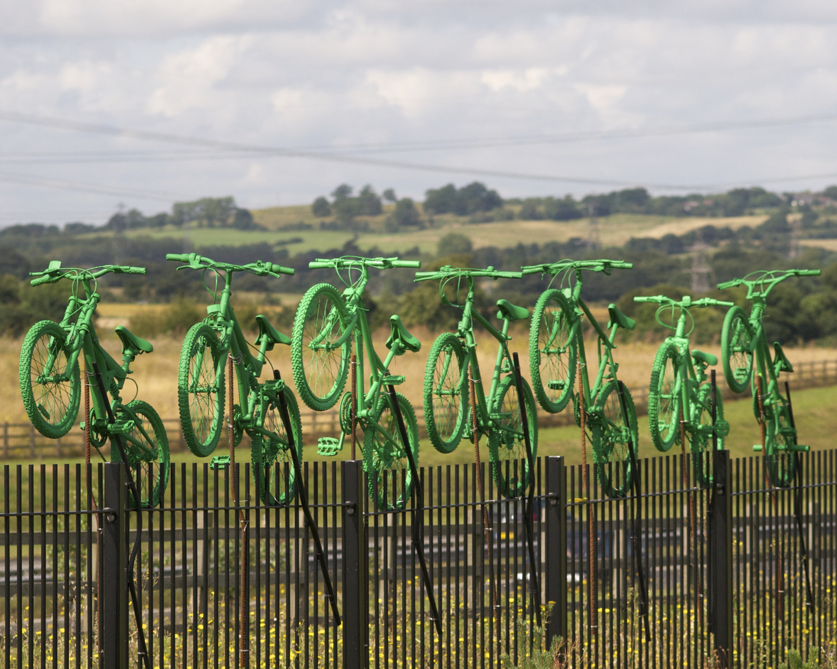 Seven green painted bikes in a line on top of the boundary fence between Cyclopark and the A2