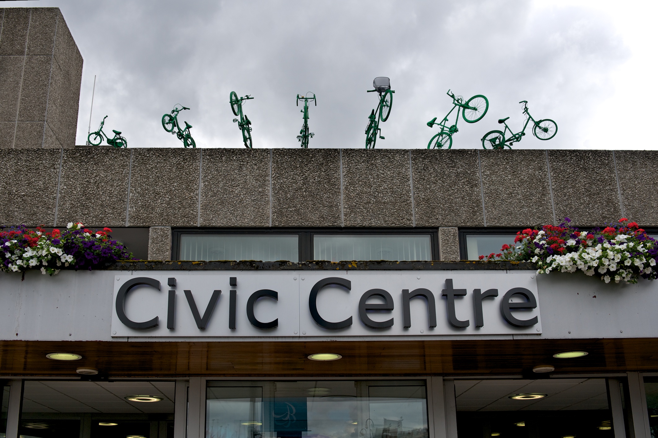 Seven green painted bikes fan out on the Civic Centre rooftop.  At one end a child's bike with stabilisers and at the other, an adult trike