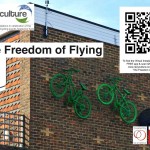 Two green bikes attached the top of a brick wall,  with title, logos & QR code