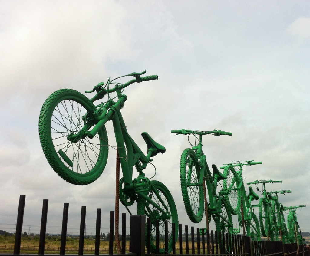 Seven green painted bikes, 'leap' from the top of the boundary fence between Cyclopark and the A2
