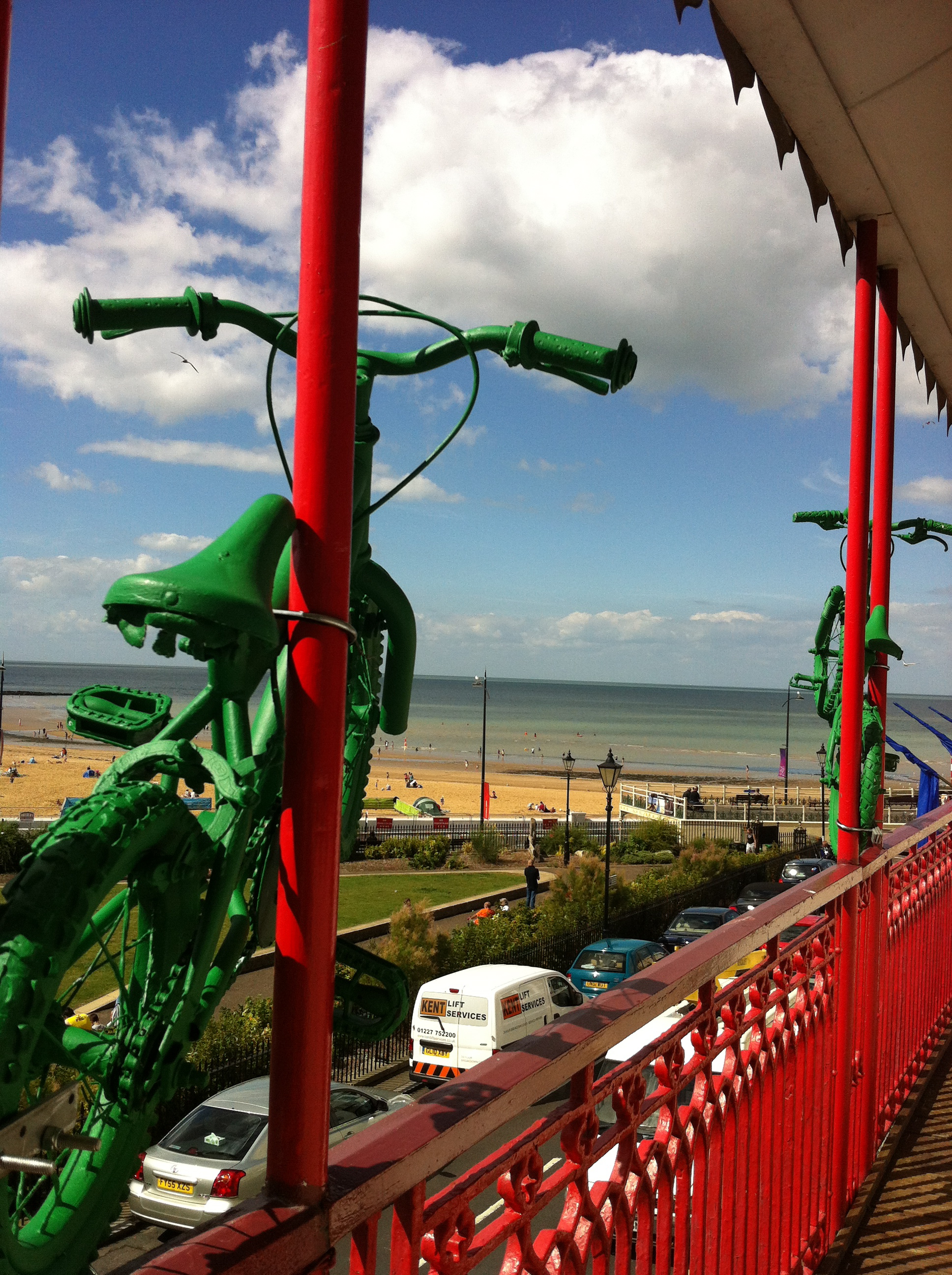 Two green painted child's bikes perched on a bright red balcony, facing out to sea