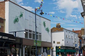 a symmetrical image of four green bikes, two on each side of the white facade of Budgens in Whitstable High St facing towards the centre
