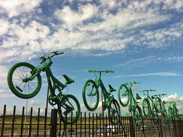 Seven green bikes atop Cyclopark's fence, overlooking the A2